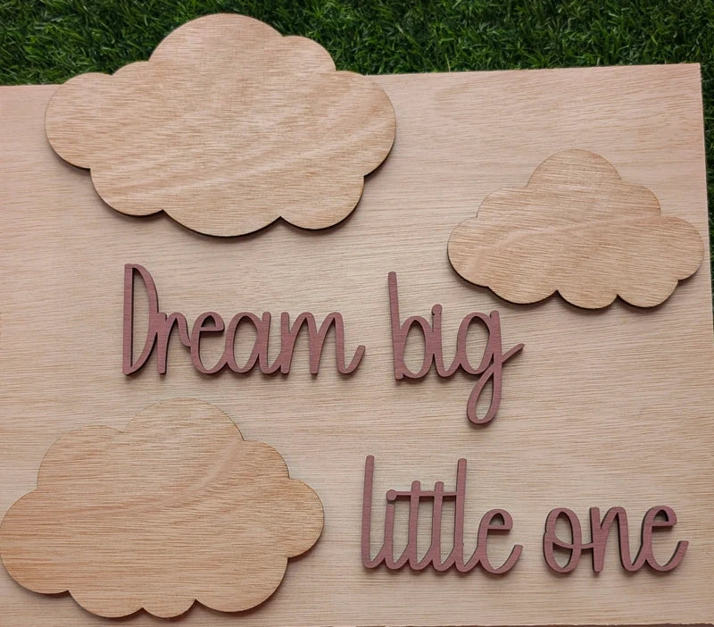 Wooden sign with 'Dream big little one' text and cloud shapes on a grass background
